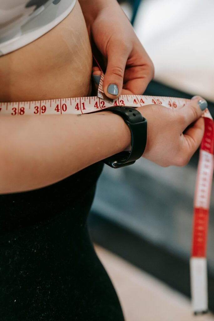 Crop anonymous female with measuring tape around big belly on blurred background of gym