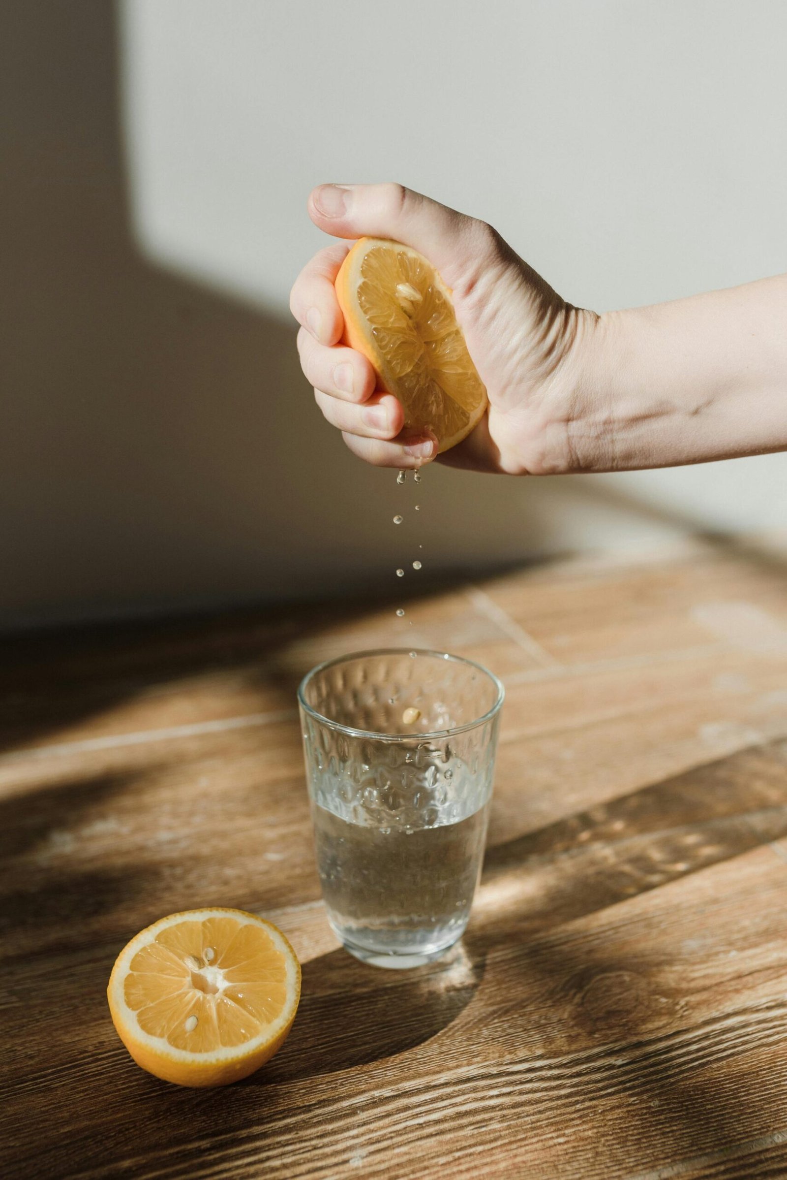 Hand squeezing a fresh lemon over a glass of water on a wooden surface, highlighting freshness.